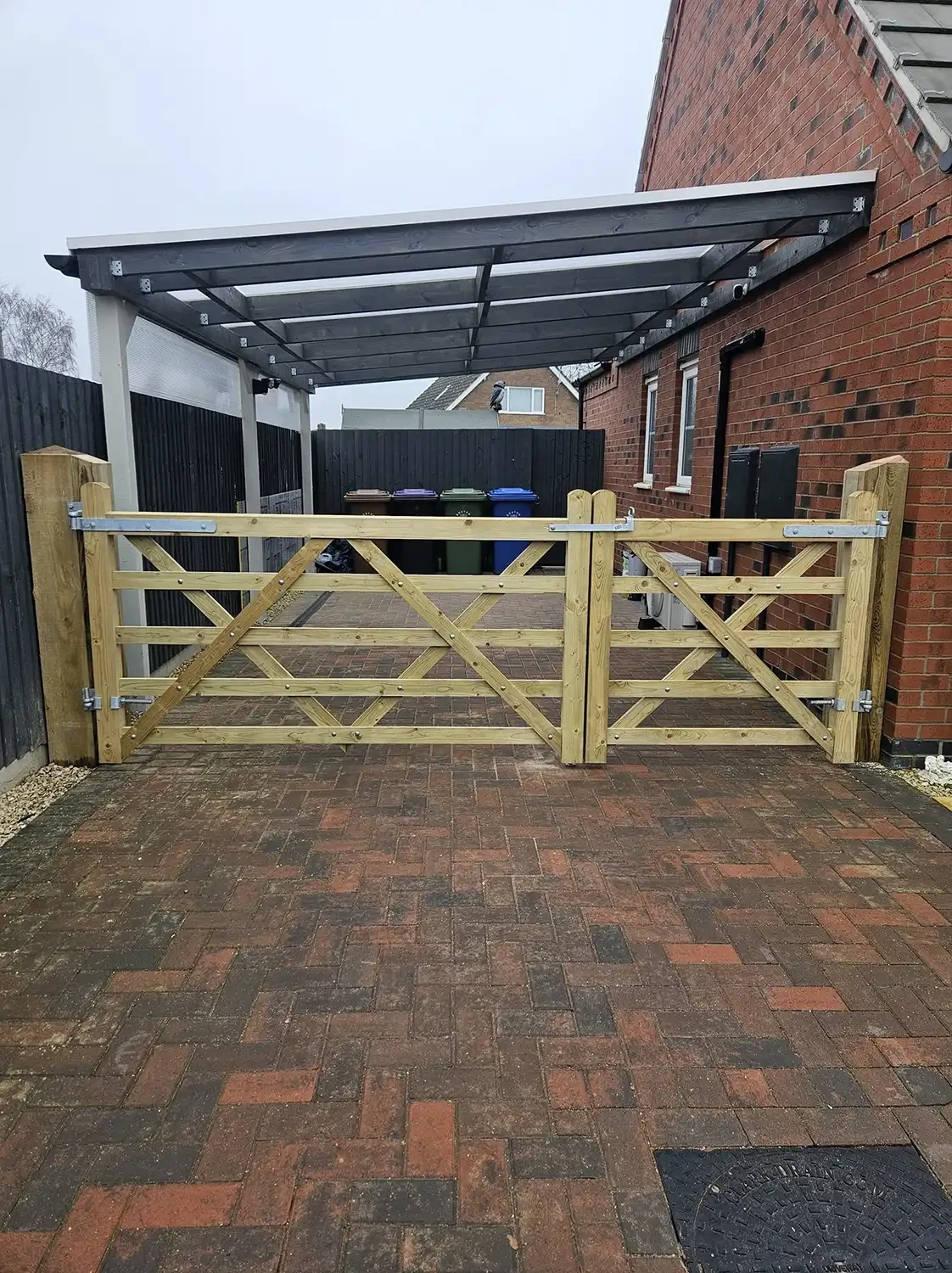 Wooden gate with diagonal slats, set on a brick-paved driveway, leading to a covered area with recycling bins in the background.
